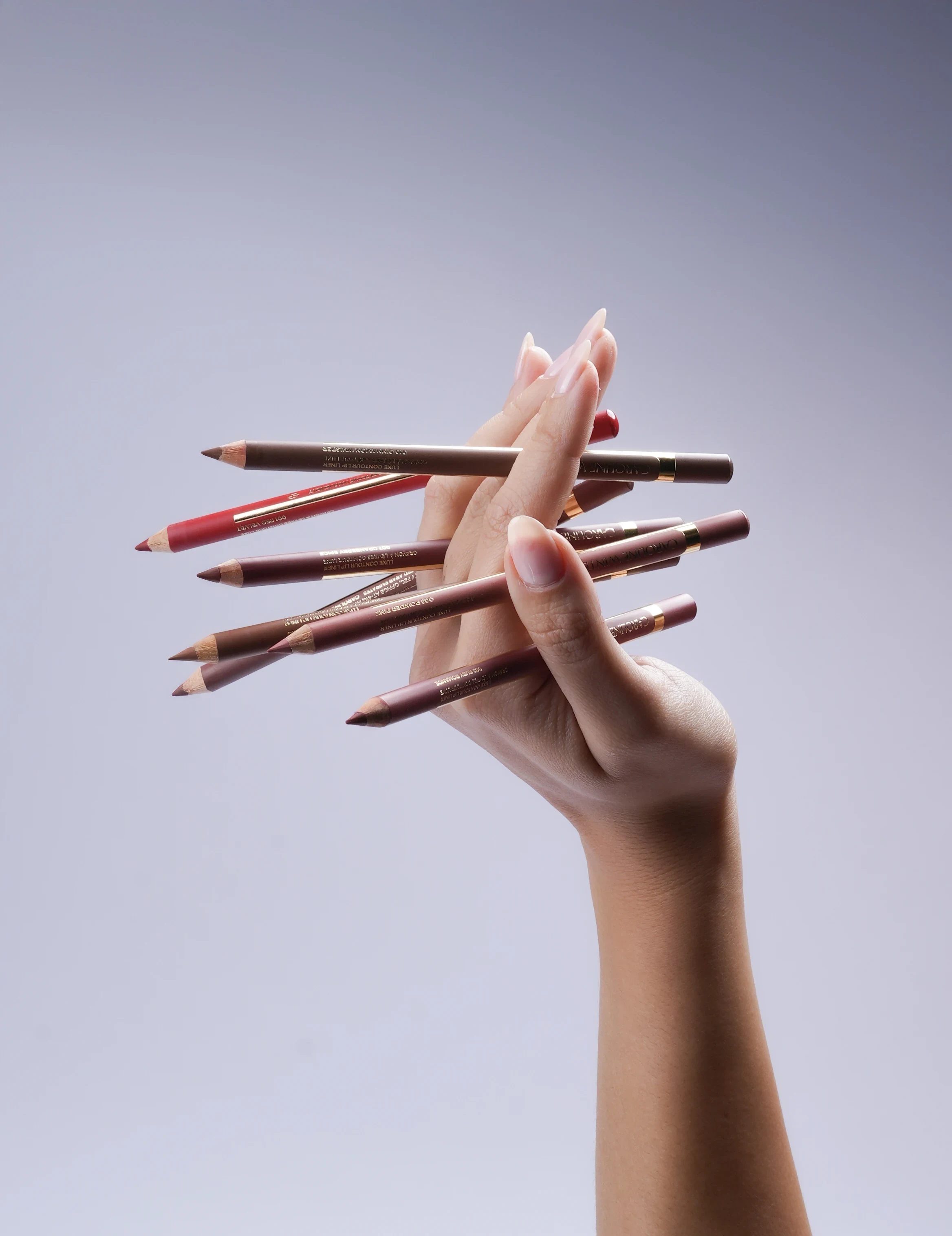 A hand holding a fan of lip liners against a soft lilac backdrop.