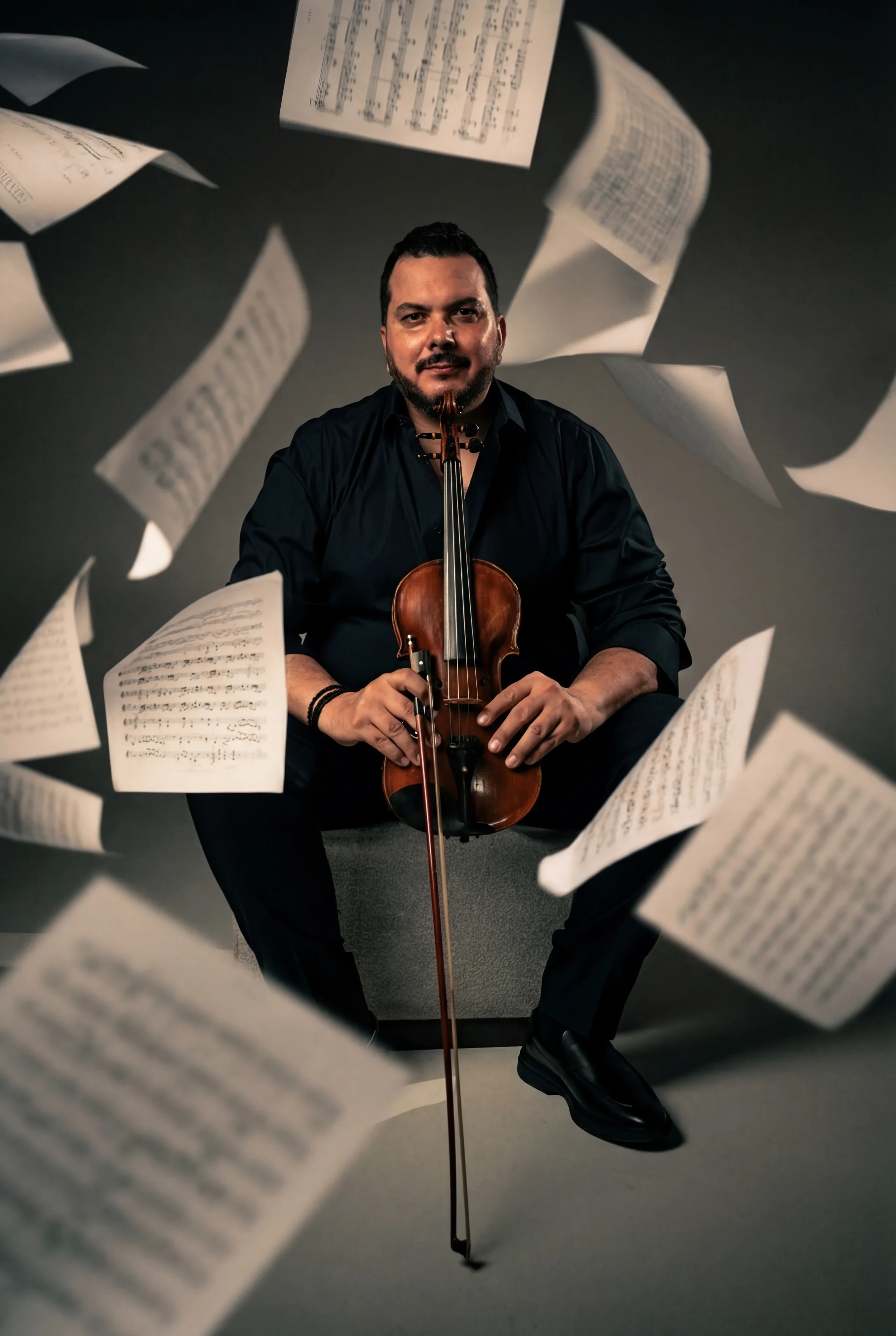 A violinist seated amid sheets of music suspended mid-air against a dark backdrop.